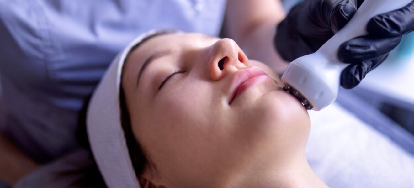A young woman in a bathrobe lies and receives a facial cleansing procedure using a device at a spa or beauty center. A natural female face with a healthy skin texture. Aesthetic cosmetology. Skincare