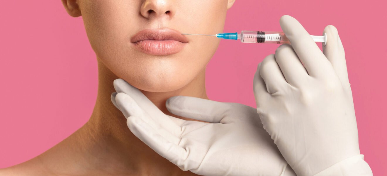 Cropped of woman receiving lip injections, focusing on her lips and the gloved hand holding the syringe. The woman is looking straight ahead, and the background is a solid pink color