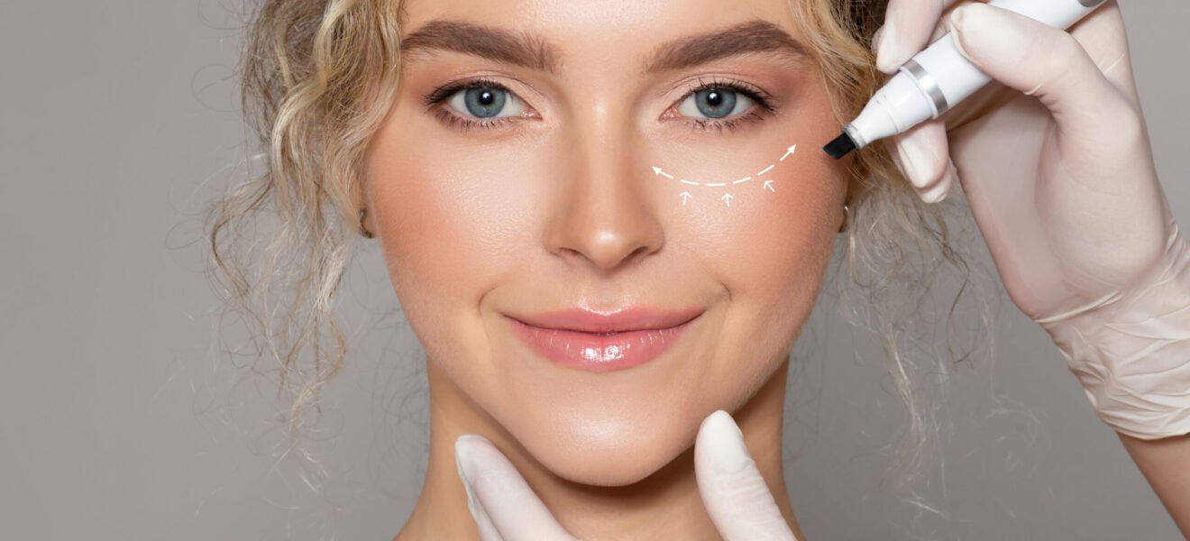 Plastic surgeon drawing contours with black pen around female eye before surgery, young woman looking at camera, isolated on grey background