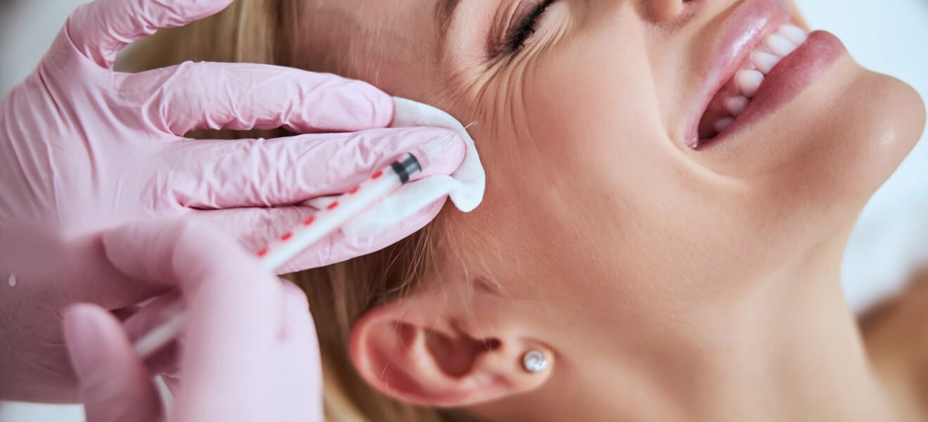 Smiling lady lying with her eyes closed during the rejuvenation injection administered by a professional dermatologist